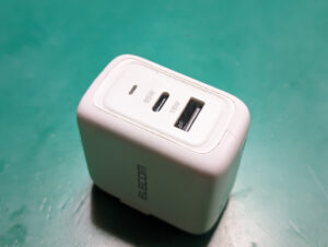 ELECOM製の白いUSB充電器。USB-Cポートは65W出力、USB-Aポートは18W出力で表示されており、緑色の背景の上に配置されている。